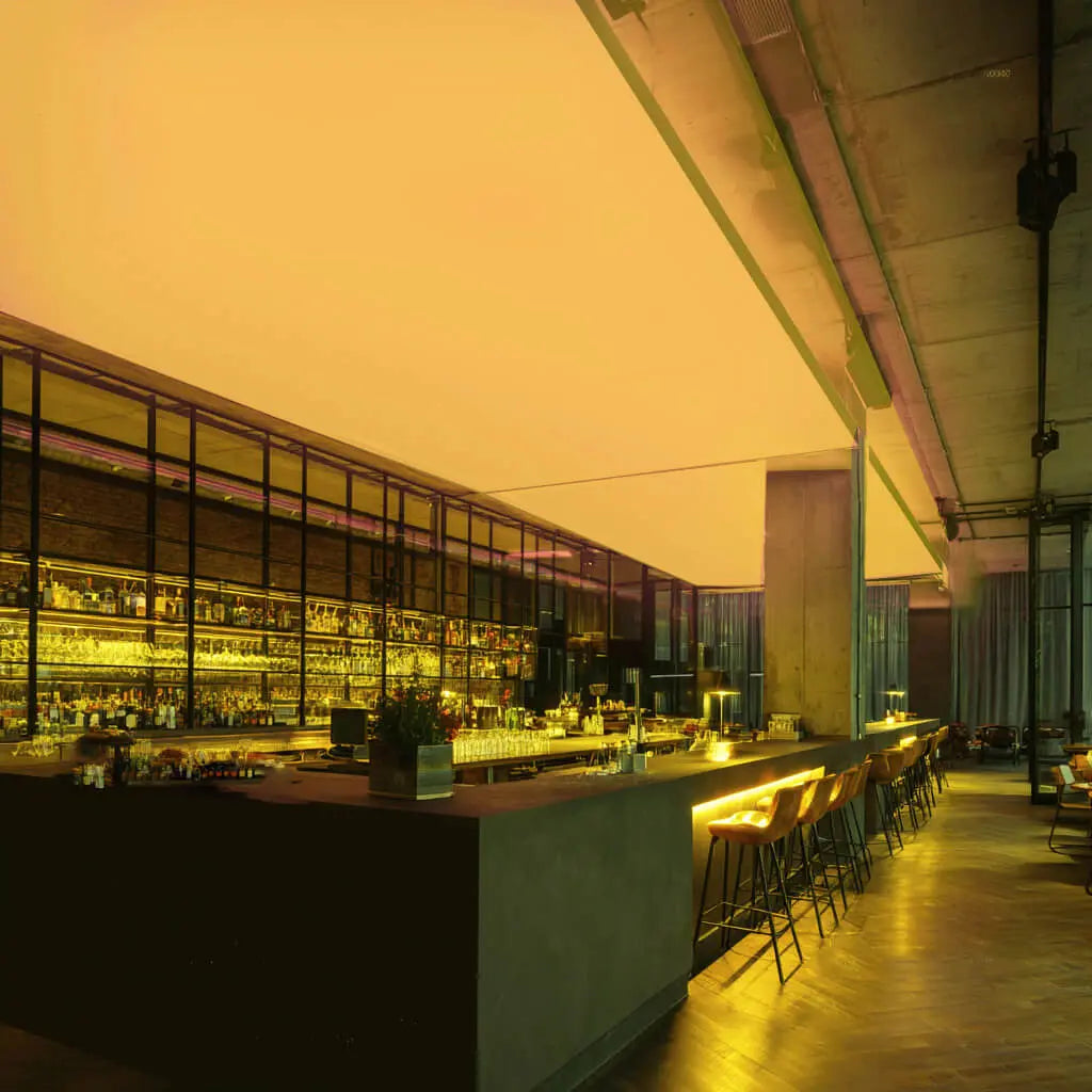 Moderne Bar mit langen Theken und Barhockern, an der Decke eine großflächige RGBW-LED-Lichtdecke, die den Raum in kräftigem Gelb beleuchtet.