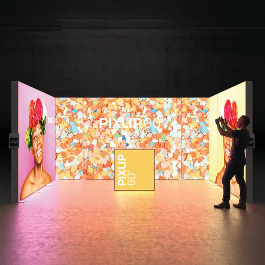 Beleuchteter PIXLIP GO Messestand mit farbenfrohen Messewänden und integrierter Theke mit Stoffbanner .