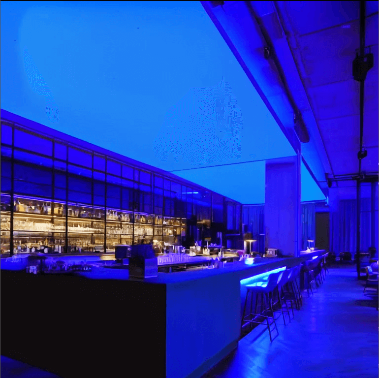 Moderne Bar mit langen Theken und Barhockern, an der Decke eine großflächige RGBW-LED-Lichtdecke, die den Raum in kräftigem Blau beleuchtet.