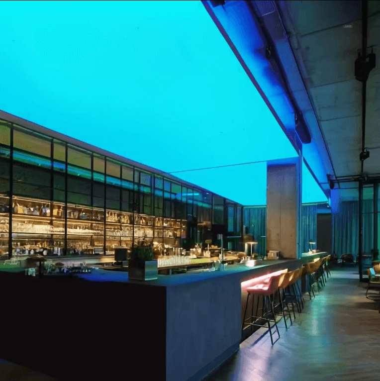 Moderne Bar mit langen Theken und Barhockern, an der Decke eine großflächige RGBW-LED-Lichtdecke, die den Raum in Hellblauer beleuchtet.