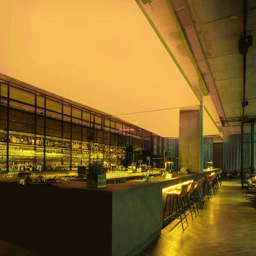 Moderne Bar mit langen Theken und Barhockern, an der Decke eine großflächige RGBW-LED-Lichtdecke, die den Raum in kräftigem Gelb beleuchtet.