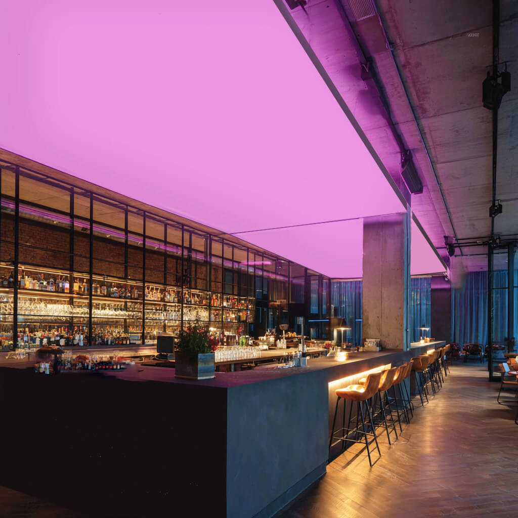 Moderne Bar mit langen Theken und Barhockern, an der Decke eine großflächige RGBW-LED-Lichtdecke, die den Raum in kräftigem Pink beleuchtet.