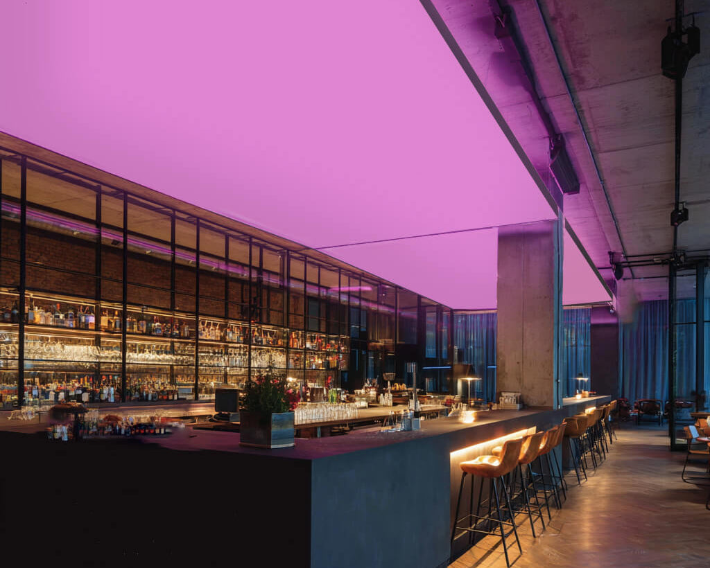 Moderne Bar mit langen Theken und Barhockern, an der Decke eine großflächige RGBW-LED-Lichtdecke, die den Raum in kräftigem Pink beleuchtet.