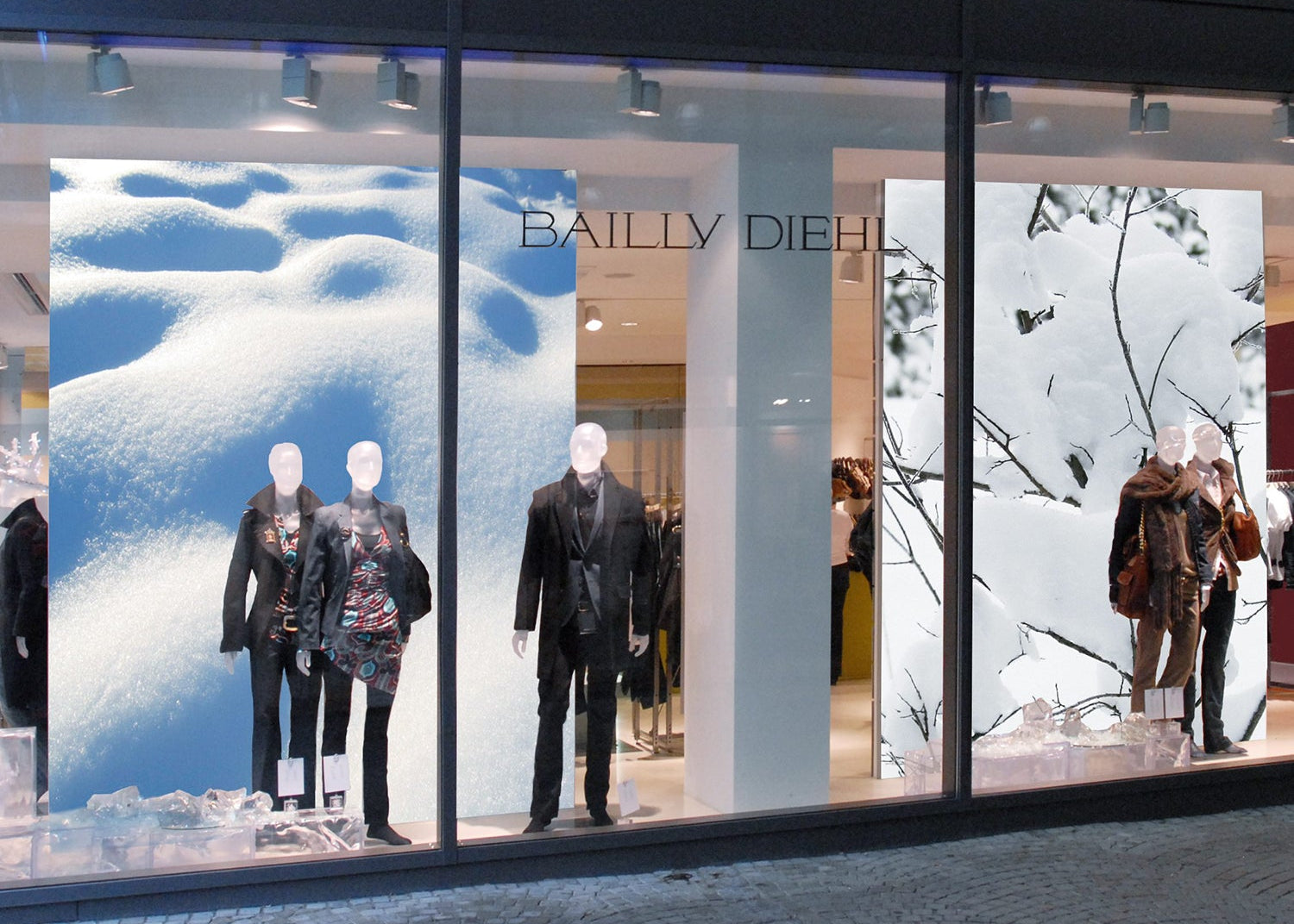 Mode-Schaufenster mit winterlicher Dekoration und großflächigen Textilspannrahmen im Hintergrund, die hochwertige Stoffdrucke mit Schneelandschaften zeigen und die präsentierte Kleidung stimmungsvoll in Szene setzen.