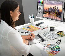 Eine Frau sitzt an einem Schreibtisch vor zwei PCs und prüft konzentriert die Grafikdaten auf den Bildschirmen.