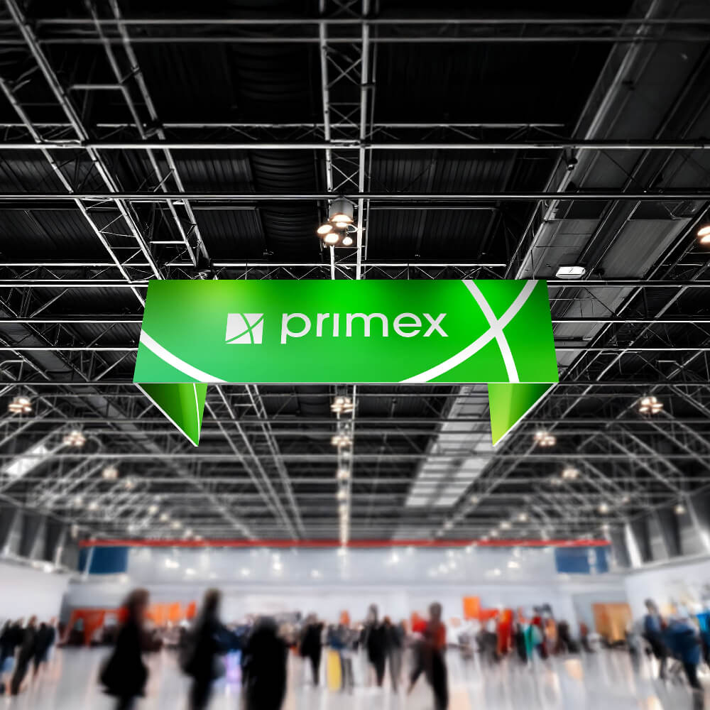 Messehalle mit einem großen, grünen, unbeleuchteten Deckenhänger in U-Form, der das weiße Logo und den Schriftzug „primex“ zeigt. Im Hintergrund sind Menschen und Messestände zu sehen.