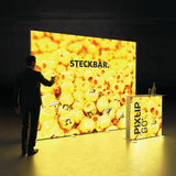 Beleuchteter PIXLIP GO Messestand mit modularer Rückwand und passender Theke in leuchtendem Gelb mit Stoffbanner .