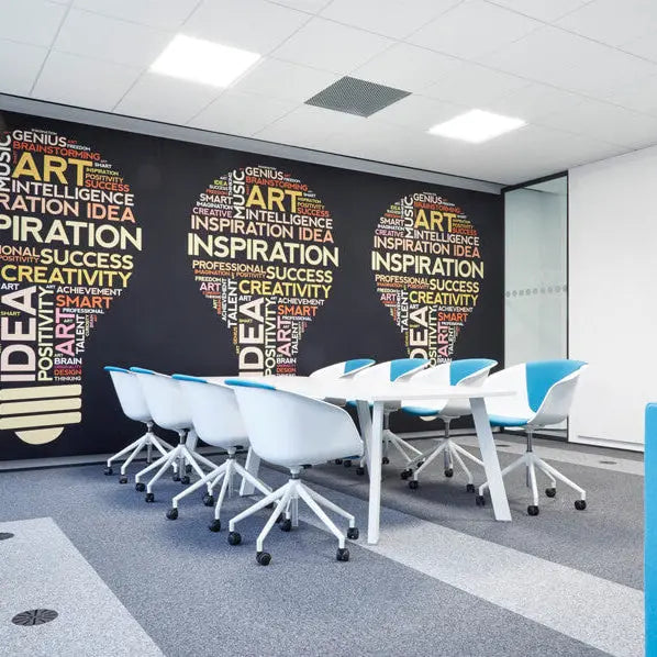 Moderner Besprechungsraum mit weißen Tischen und blauen Stühlen, an der Wand ein Akustikbild mit  drei große Glühbirnen-Grafiken aus bunten Wortwolken mit Begriffen wie Inspiration, Idee und Kreativität.